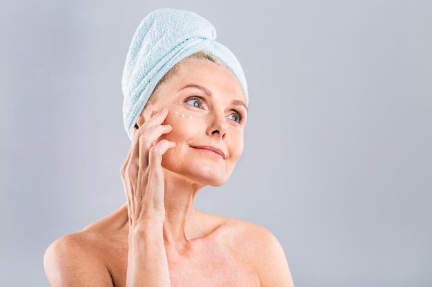 sênior sorrindo 50 anos, mulher madura de meia-idade aplicando creme facial no rosto, olhando para a câmera, anti-idade, pele seca saudável, conceito de terapia de beleza, tratamento de pele antigo
