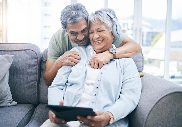 Senior umarmt sich und ein Paar mit einem Tablet für eine Website, die Musik im heimischen Wohnzimmer streamt, lachendes Sofa und ein älterer Mann und eine ältere Frau mit einem Film oder einem lustigen Video über Technologie oder gemeinsames Zuhören
