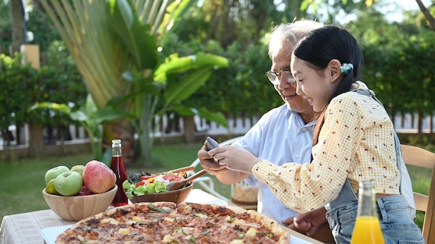 Senior und Enkelin essen Pizza im Garten zu Hause. Lebensstil im Ruhestandsalter mit der Familie im Sommerurlaub.