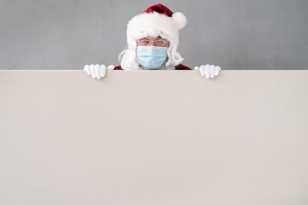 Senior vistiendo traje de Santa Claus y máscara protectora. Hombre que sostiene el tablero en blanco vacío. Vacaciones de Navidad durante el concepto COVID 19 de coronavirus pandémico