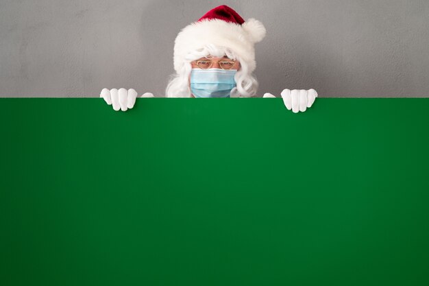 Senior vistiendo traje de Santa Claus y máscara protectora. Hombre que sostiene el tablero en blanco vacío verde. Vacaciones de Navidad durante el concepto COVID 19 de coronavirus pandémico