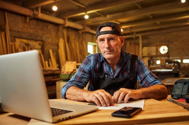 Senior Zimmermann arbeitet am Computer und schaut in die Kamera in der Werkstatt