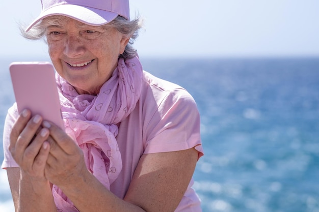Seniorin, attraktive Frau, rosa gekleidet, sitzt in der Nähe des Meeres an sonnigen Tagen mit dem Handy