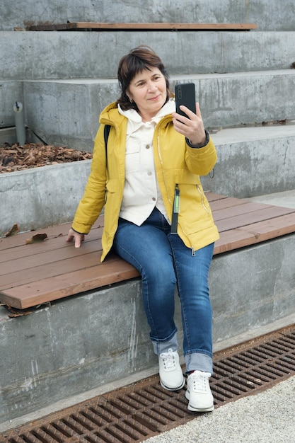 Seniorin, brünette Frau sitzt in einem Stadtpark auf den Stufen und lächelt, während sie über eine Videoverbindung spricht