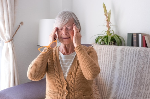 Seniorin in Freizeitkleidung sitzt zu Hause auf dem Sofa mit den Händen an den Schläfen wegen Schmerzkopfschmerzen