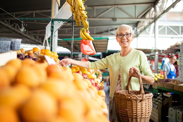 Seniorin kauft frisches Gemüse und Obst auf dem Marktplatz und hält eine Tasche voller gesunder Lebensmittel