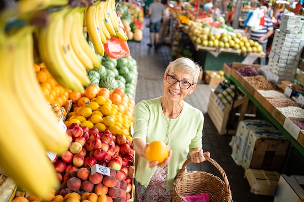 Seniorin kauft frisches Gemüse und Obst auf dem Marktplatz und hält eine Tasche voller gesunder Lebensmittel