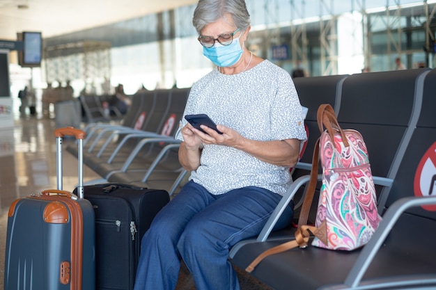 Seniorin mit Gesichtsmaske, die mit Gepäck am Flughafen sitzt und das Telefon benutzt, das auf den Abflug wartet