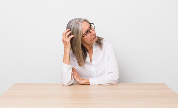 Seniorin mit grauen Haaren, die sich verwirrt und verwirrt fühlt, wenn sie den Schreibtisch und das Geschäftskonzept kratzt