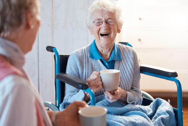 Seniorin und Kaffee oder Tee im Rollstuhl wegen einer Behinderung oder Rehabilitation und glücklich oder lachend über lustige Gespräche oder Witze Alte Menschen zusammen zum Spaß im Pflegeheim