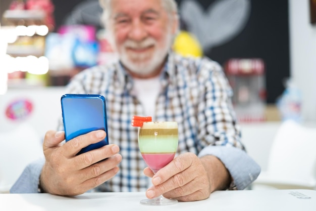 Senioro hombre usando teléfono sentado cerca de un vaso de especialidad local de la isla canaria llamado barraquito hecho con leche condensada, café y licor