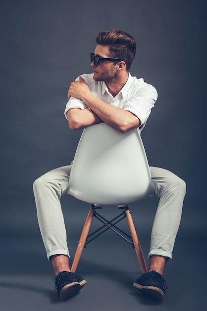 Señor Style. Longitud total de hombre guapo joven confiado en gafas de sol mirando a otro lado mientras está sentado en una silla contra el fondo gris