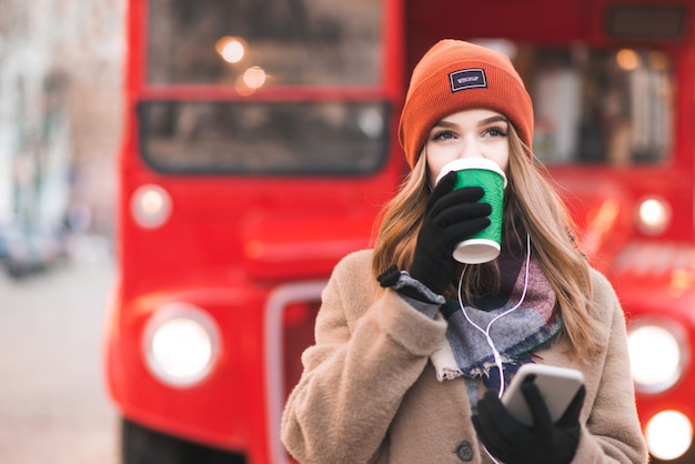 Señora con un abrigo y auriculares está de pie sobre un fondo rojo de la ciudad con un teléfono inteligente en sus manos, tomando café de una taza verde y mirando a otro lado