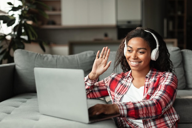 Señora afroamericana milenaria sonriente en auriculares inalámbricos que estudian con la mano que agita de la computadora portátil