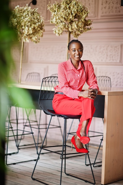 Señora afroamericana de negocios de trenzas bonitas brillante persona mandona amigable usar camisa roja de oficina y pantalones, sentado en la silla.