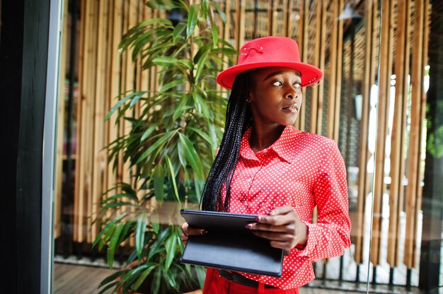 Señora afroamericana de negocios de trenzas bonitas brillante persona mandona ropa de oficina camisa roja, sombrero y pantalones, mantenga la tableta en las manos.
