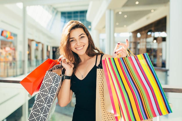 Señora alegre en elegante vestido negro llevando bolsas de colores mientras camina en la tienda