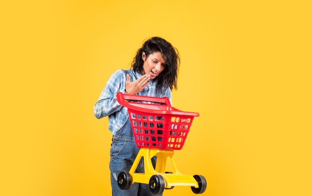 Señora alegre en estilo de mezclilla de camisa llevar carrito de compras en compras de fondo amarillo