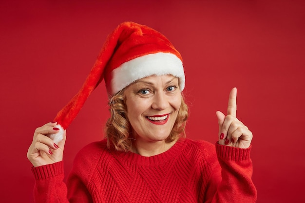 Señora alegre en una gorra de año nuevo posa sobre un fondo rojo, sostiene el dedo índice hacia arriba