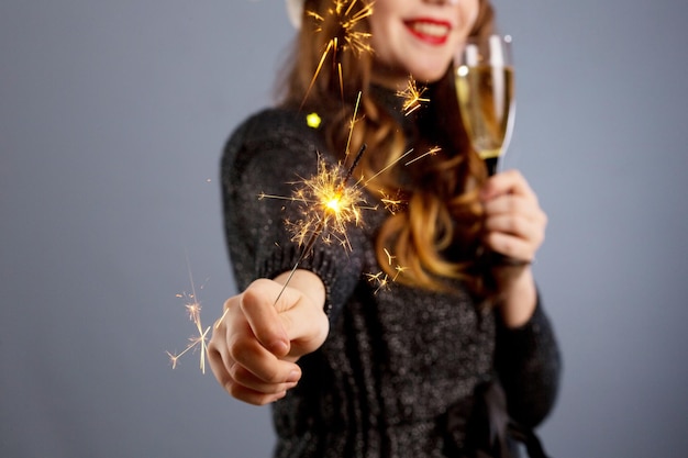 Señora alegre con el pelo rizado en el sombrero de Navidad sonriendo sosteniendo una bengala y bebiendo champán