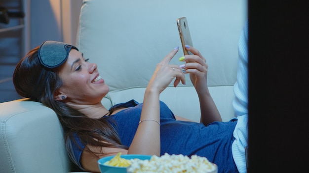 Señora con antifaz para dormir en la frente y pijamas buscando en el teléfono acostado en el sofá. Mujer feliz sentada en el sofá leyendo, buscando, navegando en el teléfono inteligente usando internet móvil