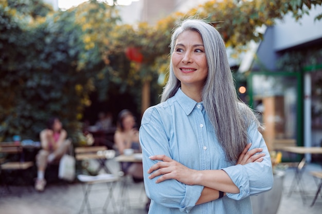 Señora asiática canosa positiva con los brazos cruzados contra la terraza de un café al aire libre