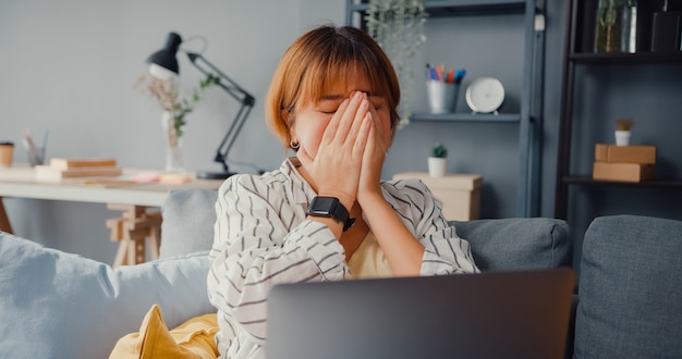 Señora asiática independiente siente dolor de cabeza mientras está sentada en el sofá con la computadora portátil en línea, aprende en la sala de estar de la casa