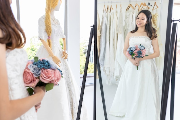 Foto señora asiática está mirando en el espejo y sonriendo mientras elige vestidos de novia en la tienda.