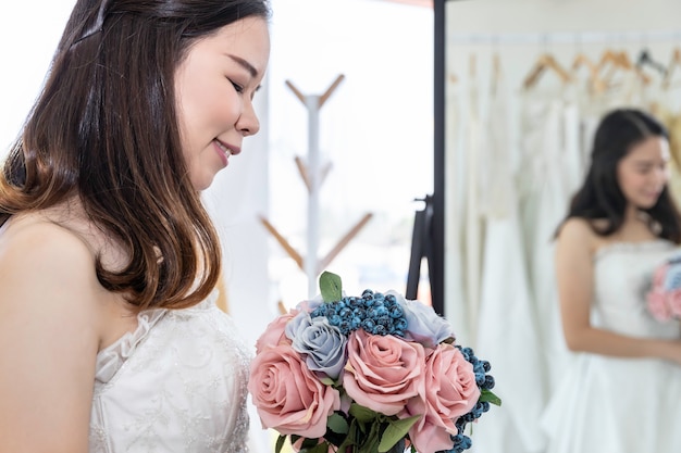 Señora asiática está mirando en el espejo y sonriendo mientras elige vestidos de novia en la tienda.