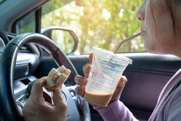 Señora asiática sosteniendo café helado y comida de panadería de pan en un automóvil peligroso y corre el riesgo de sufrir un accidente