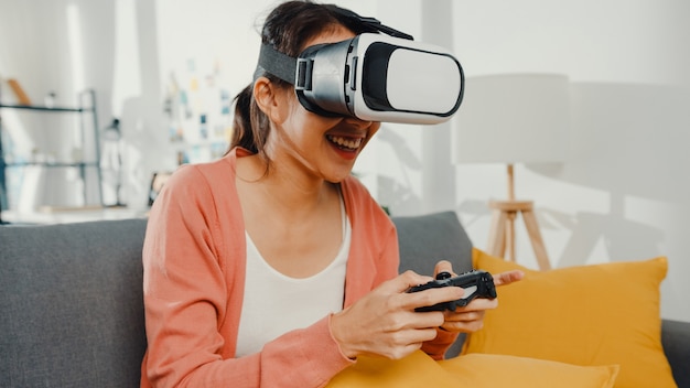 Señora asiática usa gafas de realidad virtual juego de joystick en el sofá en el salón de la casa