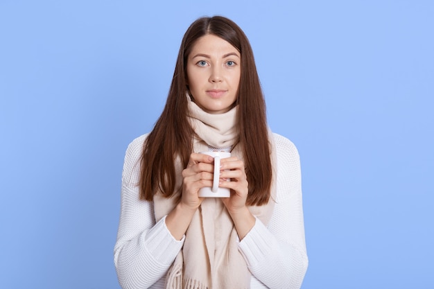 Señora de aspecto agradable con jersey y bufanda posando aislado sobre fondo azul con taza de té en las manos