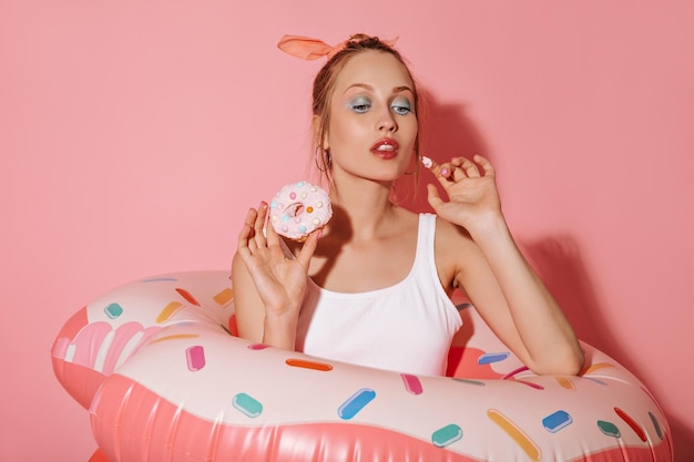 Señora atractiva con maquillaje brillante en traje de baño blanco sosteniendo donut y posando con grandes anillos de natación sobre fondo rosa