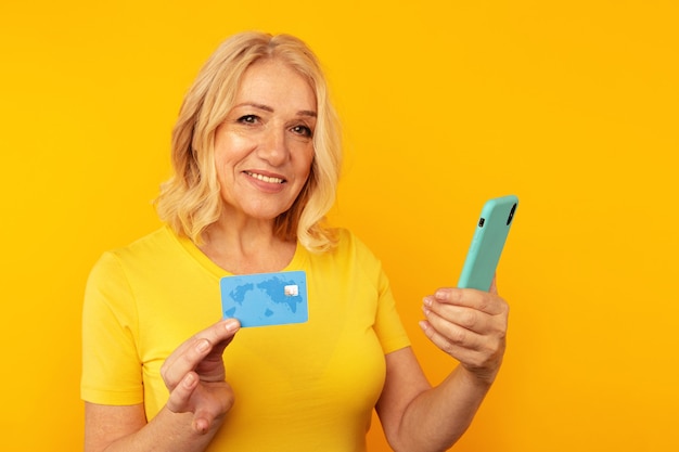 Señora bastante adulta en ropa brillante mirando la cámara y posando con teléfono azul y tarjeta de crédito.