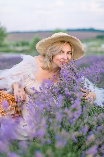 Foto señora bastante madura con elegante sombrero y vestido, agachándose y disfrutando del aroma de la lavanda en flor