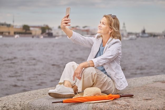 La señora blanca atractiva activa mayor de unos 60 años está sentada cerca del río en un paño informal y elegante con el teléfono móvil en las manos.
