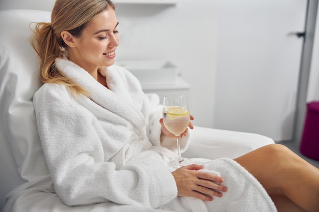 Señora bonita feliz está sentada en bata de baño en la silla de pedicura y disfrutando de un vaso de agua fría con limón