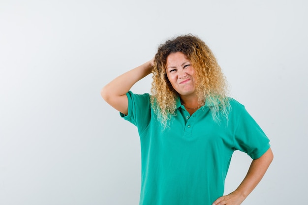 Señora bonita rubia en camiseta de polo verde rascándose la cabeza mientras frunce el ceño y mira olvidadiza, vista frontal.