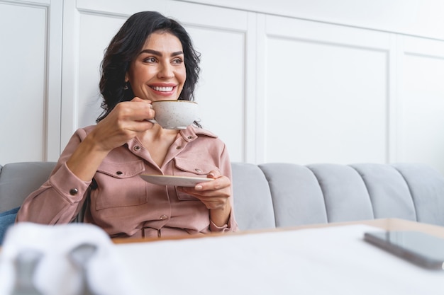 Señora bonita sonriente sentada en la mesa de café y disfrutando de una deliciosa bebida caliente