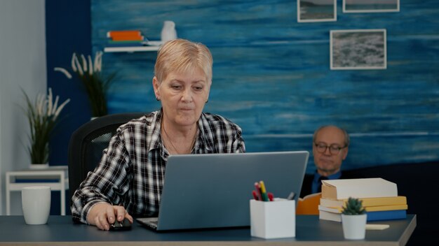 Señora caucásica madura trabajando en la computadora portátil en el lugar de trabajo en casa, escribiendo en la computadora tomando café. Gerente experimentado haciendo proyecto financiero durante el autoaislamiento mientras el esposo lee un libro en backgro