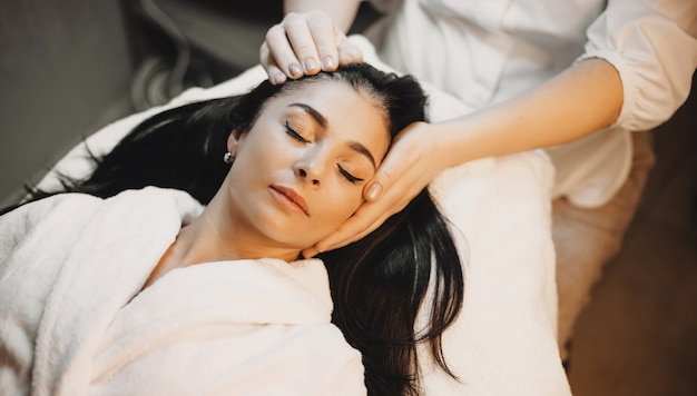 Señora caucásica morena está mintiendo y teniendo un procedimiento de masaje de cabeza en un salón de spa profesional