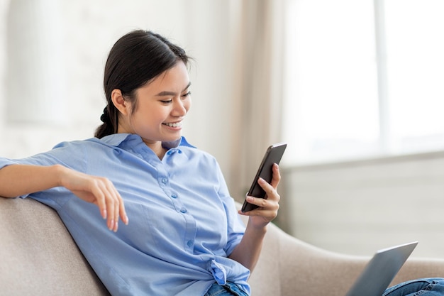 Señora china feliz que usa la computadora portátil y el teléfono inteligente en casa