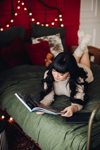 Señora descansando en una cama, decorada con luces de cadena, y mirando un libro en sus manos.