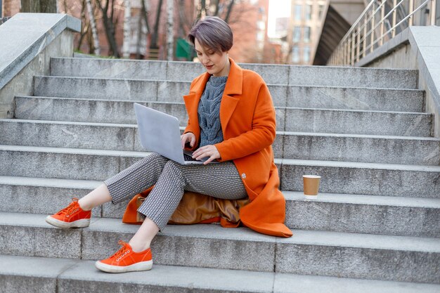 Señora elegante en abrigo naranja brillante con su computadora portátil y taza de café