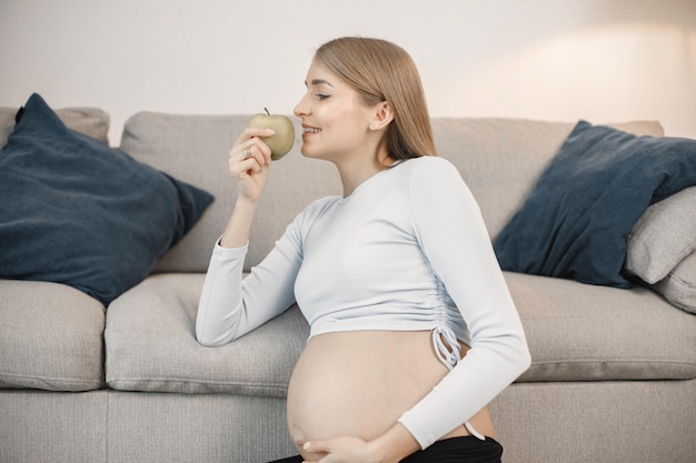 Señora embarazada sentada cerca de un sofá en la sala de estar sosteniendo una manzana
