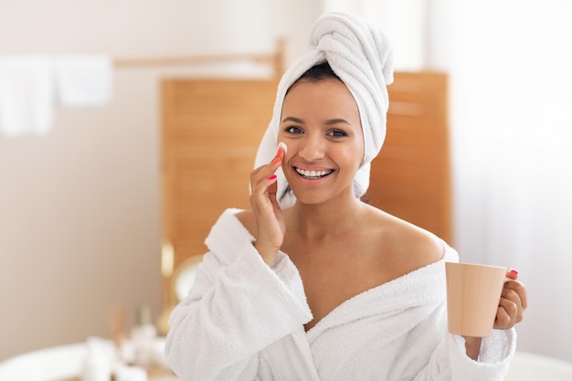 Señora feliz aplicando crema hidratante facial con taza de café en el baño