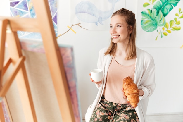 Señora feliz pintor tomando café comiendo croissant