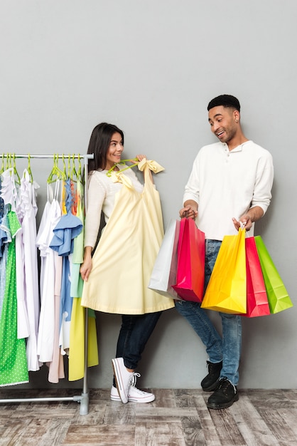 Señora feliz que elige la ropa cerca del hombre que sostiene bolsos de compras.