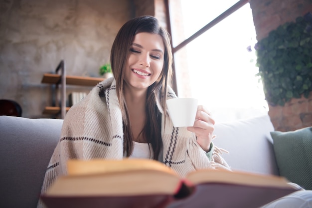 Señora hogareña beber café leer un libro novedoso sentarse sofá manta caliente cubierta en la sala de estar