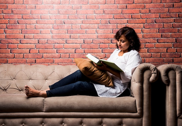 Señora india de mediana edad o mujer leyendo un libro mientras está sentado en un sofá o sofá, tomando un café